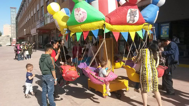 新疆の対照的なイメージ: 回転木馬に乗って遊ぶ子供たちの後ろでは、ホータン市場の「自警団」が訓練している。